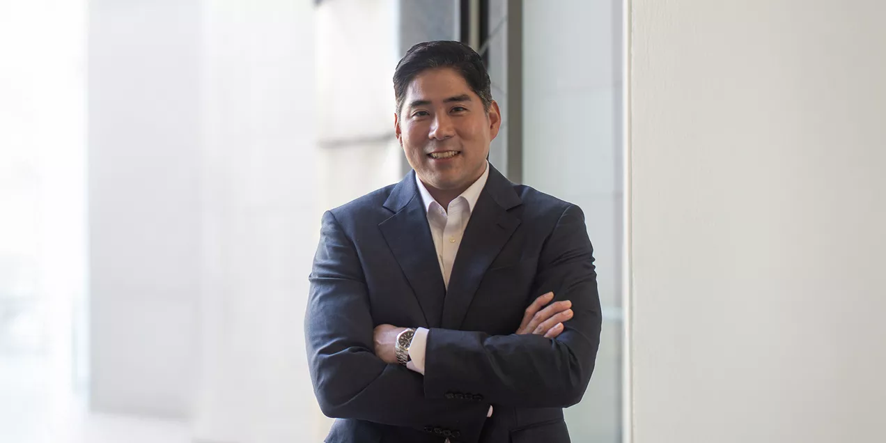 Neurosurgeon in a navy suit and white shirt stands with arms crossed, smiling in a bright hospital corridor.