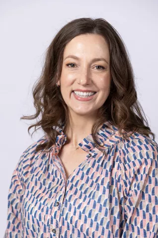 Smiling person with shoulder-length wavy brown hair, wearing a patterned blouse against a light gray background.