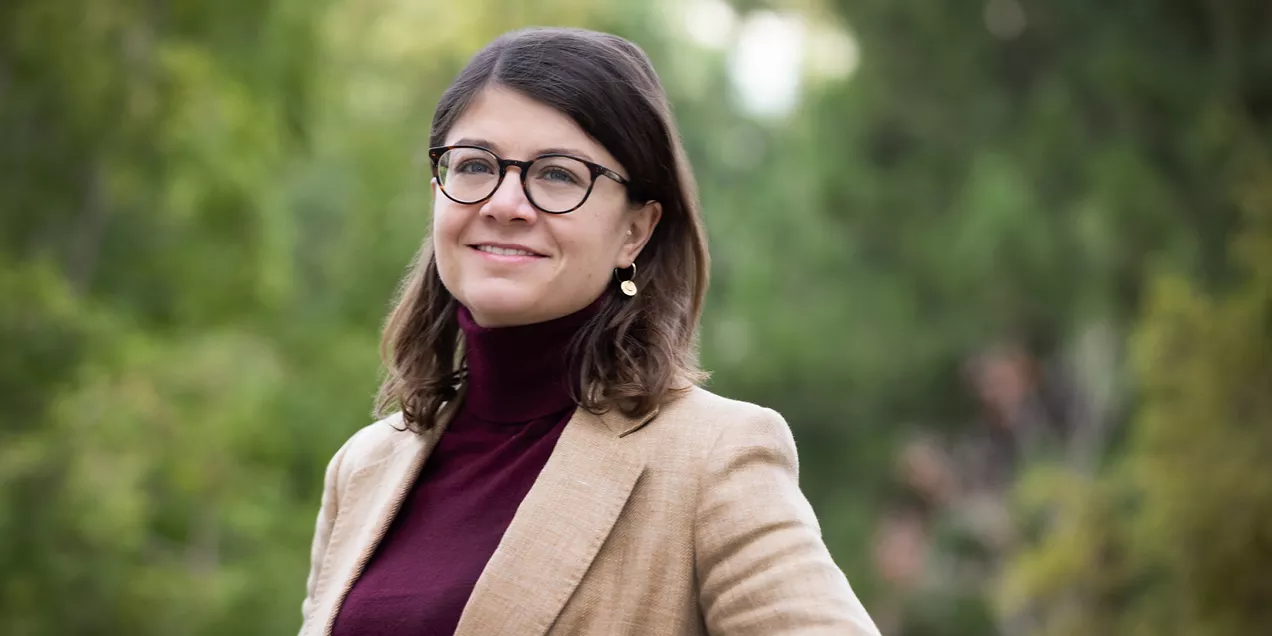 Portrait of a woman wearing glasses and a tan blazer outdoors against a softly blurred green background.
