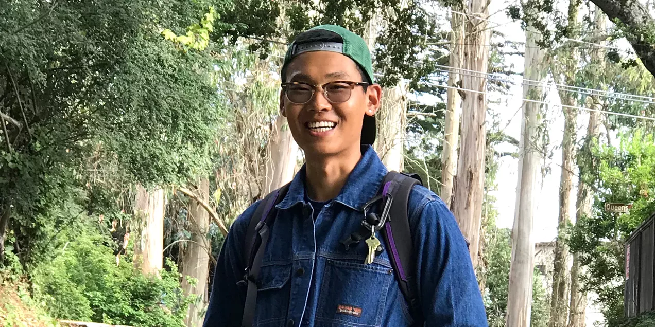 Portrait of a smiling person wearing glasses, a backward green cap, denim jacket, and backpack on a tree-lined street.
