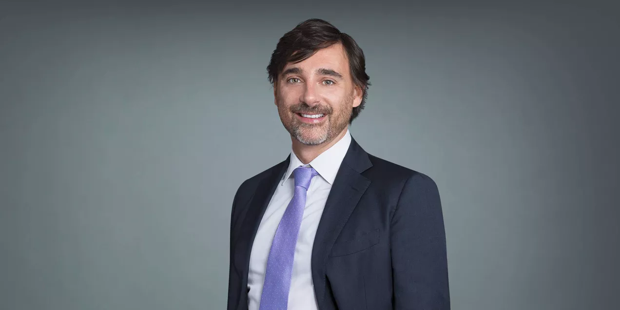 Professional headshot of Riccardo Lattanzi wearing a dark suit and lavender tie, smiling against a gray background.
