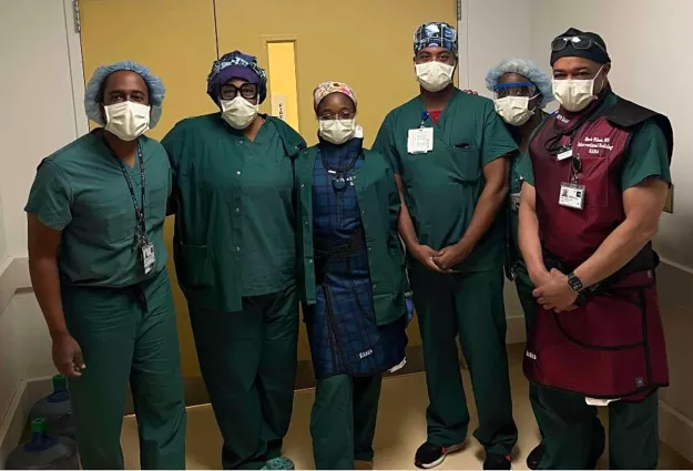Six healthcare professionals wearing surgical scrubs, masks, and protective caps stand together in a hospital hallway outside an operating room. One team member is wearing a lead apron, and the group is facing the camera for a team photo.