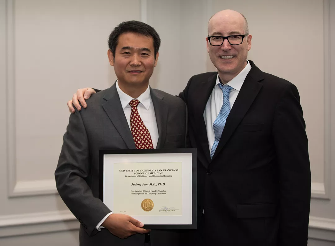 Two people in suits standing together at an award event, smiling and holding a framed certificate. One has an arm around the other in a congratulatory pose, and the setting appears to be an indoor formal venue.