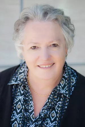 Portrait of a smiling older woman with short gray hair, wearing a patterned blouse and dark cardigan, against a soft neutral background.